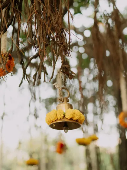 Attention to detail is key. Here, a traditional brass temple bell is adorned with fresh marigolds, hanging from the natural roots of a banyan tree to create a rustic and sacred ambiance.