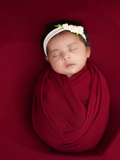 A newborn wrapped in a red swaddle, sleeping peacefully against a matching red background. The heart-shaped shadow effect adds a subtle, artistic touch of love.