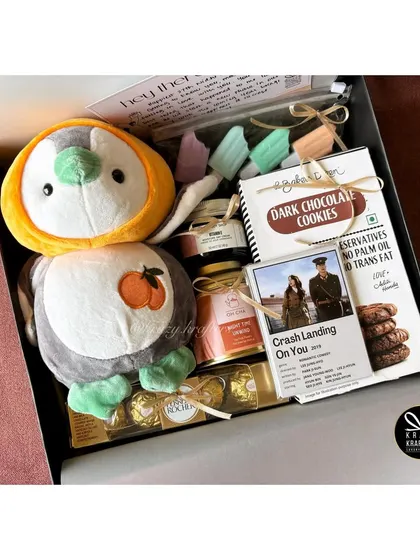A birthday hamper for a K-Drama fan, including a 'Crash Landing on You' polaroid, dark chocolate cookies, and an adorable penguin plushie.