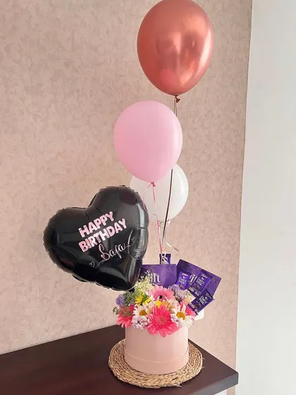 A bright and cheerful birthday hamper. The pink box is filled with fresh gerberas and other flowers, Dairy Milk chocolates, and a fun balloon bunch.