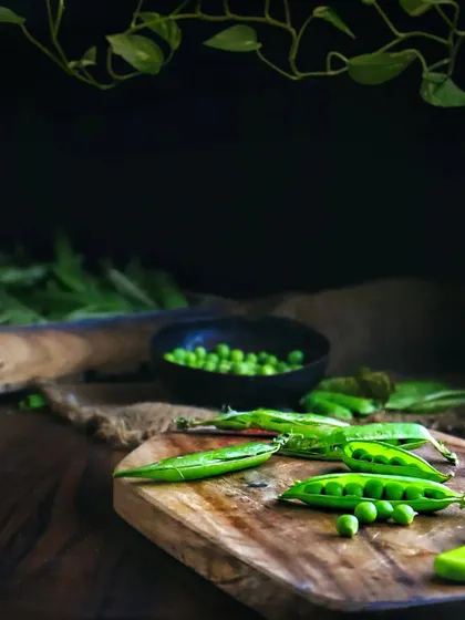 A dark and moody shot of fresh green peas on a wooden cutting board. The dramatic lighting and composition give the simple vegetable a sophisticated and artistic look.
