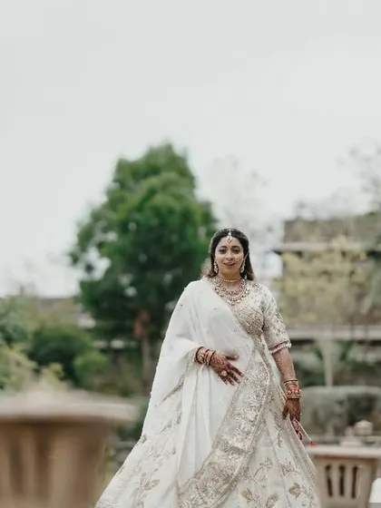 A graceful portrait of the bride in her white and gold lehenga, walking through the serene, green pathways of the wedding resort.