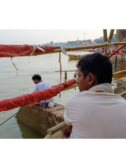 Two men sit by the edge of the Ganga in Varanasi, looking out at the water. The over-the-shoulder perspective creates an intimate and contemplative mood.