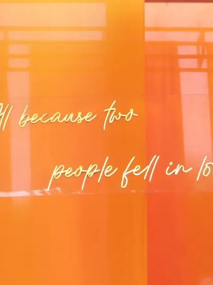 A neon sign with the quote 'All because two people fell in love' glows against a vibrant orange backdrop, adding a romantic touch.