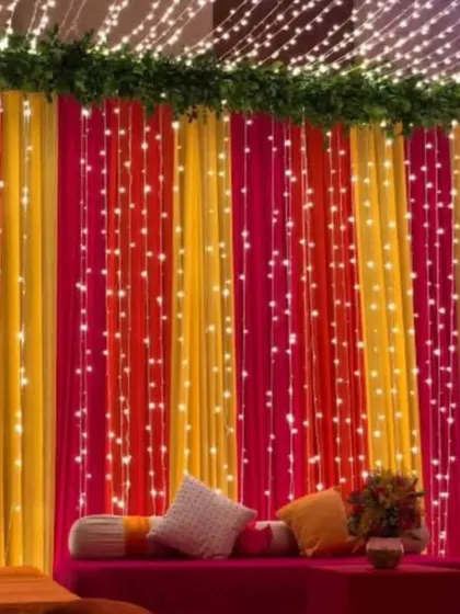 A colorful Mehendi or Sangeet backdrop combining bright yellow, pink, and orange drapes. The setup is illuminated with cascading fairy lights and features comfortable floor seating with cushions, creating a warm and inviting atmosphere.