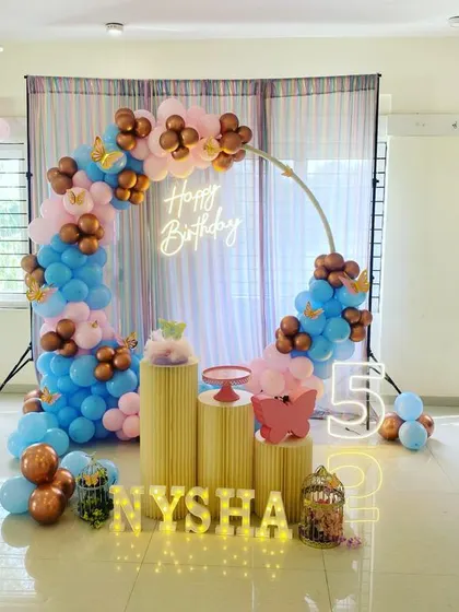 A fifth birthday party with a circular balloon arch in blue, pink, and rose gold. The setup includes gold plinths, a neon sign, and marquee letters and numbers.