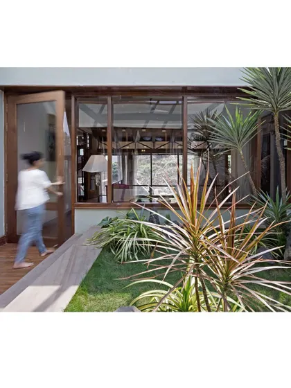 The second courtyard, seen here with a resident moving through the space, connects the lounge to the bedrooms. This open-air corridor is planted with local flora like the bird of paradise plant, enhancing the home's tropical modernism.