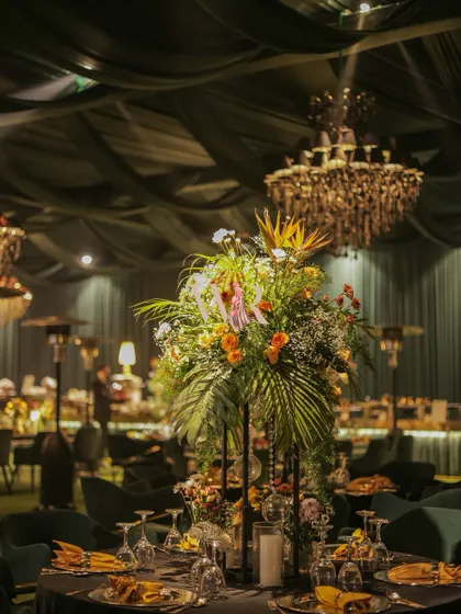 A tall, vibrant floral arrangement stands as a centerpiece on a guest table. The mix of tropical leaves and colorful flowers adds a touch of the exotic to the moody, chandelier-lit reception space.