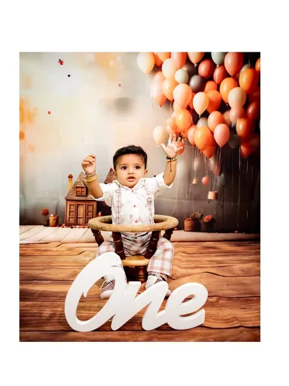 A whimsical studio portrait of a baby boy celebrating his first birthday, with a "ONE" sign and a backdrop of floating balloons.