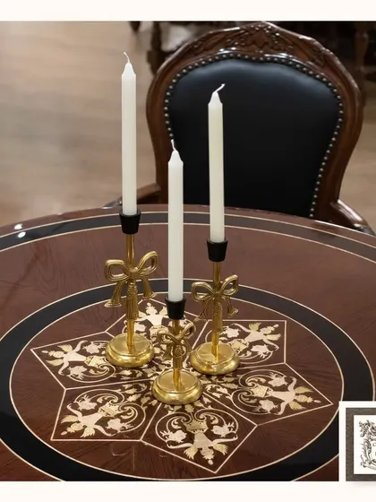 These exquisite gold-finished metal candle stands, with their delicate bow details, sit atop a dining table with a beautiful floral inlay. They are perfect for adding a warm glow to a vintage or regal interior.