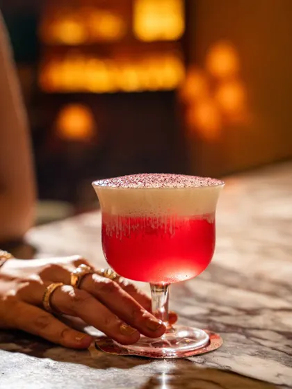 A hand gently touching a bright pink cocktail at Lyla Bombay. The intimate framing and moody lighting create a sensual and alluring image.