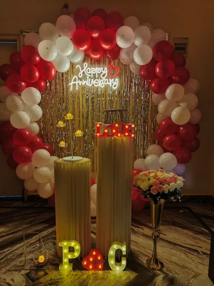 A classic anniversary decoration with a red and white balloon ring and a silver fringe backdrop. The 'Happy Anniversary' neon sign adds a warm glow to the celebration.