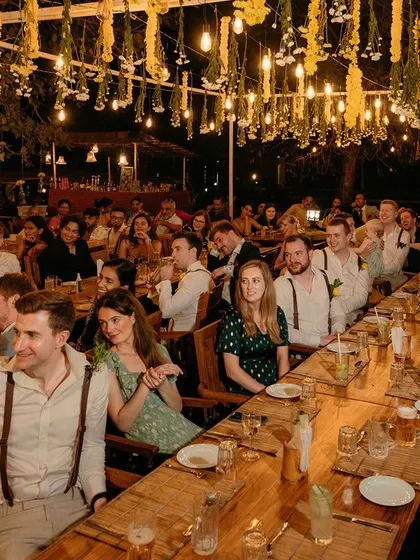 A long-table dinner setup for a rustic and intimate reception, with guests gathered under a canopy of hanging flowers and lights.