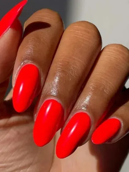 A close-up of a vibrant, almost neon-red gel polish on long almond nails, showing off the intense color and shine.