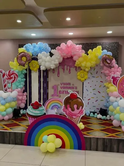 A sweet Candyland-themed first birthday. The backdrop is decorated with balloon clouds and candy props like ice cream cones, donuts, and lollipops.