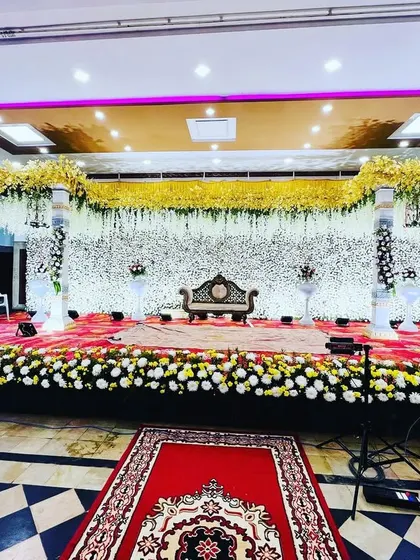 A wide shot of a beautiful reception stage with a full white floral backdrop. The design is accented with yellow flowers on the stage border and pillars, creating a fresh and elegant look.