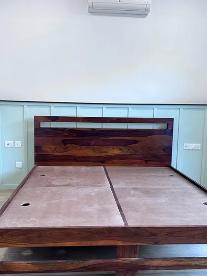 The solid wood frame of a bed, ready for the mattress. The image shows the quality of the wood and the panelled headboard design against a soft green wall.