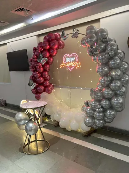 An anniversary photo point featuring a circular frame decorated with maroon, white, and silver balloons. The 'Happy Anniversary' neon sign and butterfly accents add a touch of romance to the design.