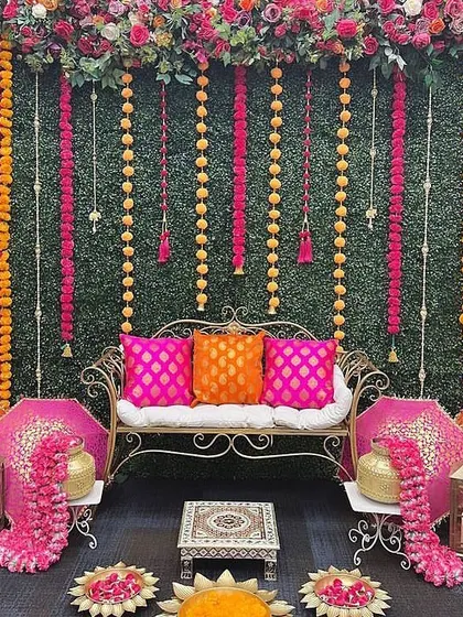 A chic Mehendi seating area with a green turf wall. The hanging marigold strings and bright pink cushions add a pop of color and traditional flair.