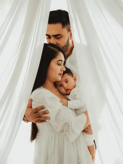 A beautifully framed family portrait, with the parents creating a loving enclosure around their baby. The sheer white curtains add a soft, dreamy quality.