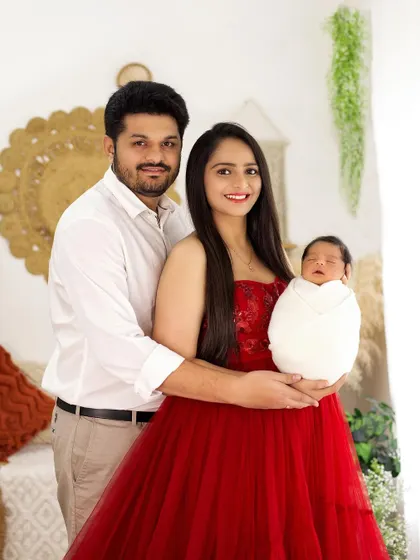 A classic family portrait with a newborn. The proud parents are smiling, holding their swaddled baby, in a warm, boho-chic studio setting.