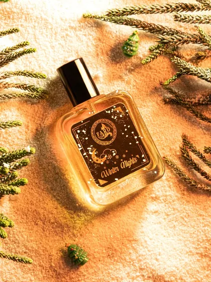 This festive flatlay for 'Winter Nights' perfume uses pine branches and a sandy texture to evoke a cozy, wintery feel. The warm, golden lighting enhances the luxurious feel of the fragrance.