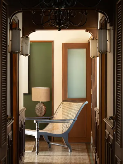 A view through a louvered doorway into the master lounge. The composition layers different spaces, textures, and light, from the cane-backed chair to the frosted glass panel, creating depth and intrigue.