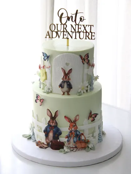 "Onto our next adventure." This charming two-tiered Peter Rabbit themed cake was created for a baby shower, celebrating the beautiful journey of welcoming a new family member.