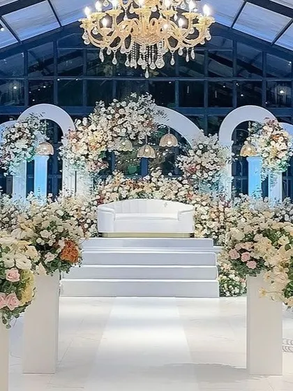 An opulent, all-white stage in a glasshouse, featuring grand arches and an abundance of flowers for a luxurious feel.