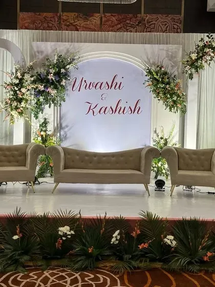 I create personalized stages that serve as a beautiful backdrop for photos. This elegant reception stage features a custom backdrop with the couple's names, flanked by sophisticated floral arrangements and comfortable lounge seating.