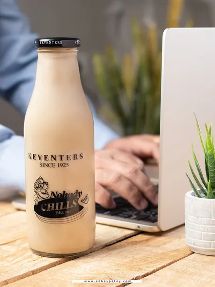 This is a lifestyle shot of a cold coffee bottle on a work desk. It places the product in a relatable, everyday context, positioning it as the perfect work-from-home refreshment.