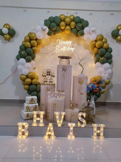 A birthday setup with a green and gold balloon ring. The decor includes multiple white plinths of varying heights, decorative birdcages, and marquee letters.