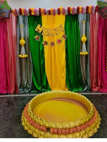 A Haldi ceremony setup with a large 'urli' and a backdrop of colorful drapes. A "Haldi" banner adds a personalized touch.
