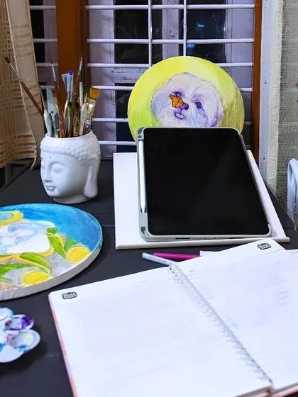 A quiet corner of my art desk. You can see my sketchbook for jotting down ideas, a Buddha head holding my brushes, and some finished pieces like a pet portrait, all part of my creative ecosystem.