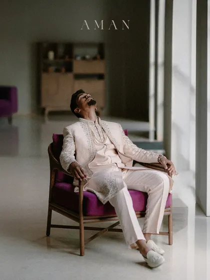 An artistic portrait of a groom leaning back in a chair, bathed in light. The pose is relaxed yet stylish, capturing a moment of calm before the festivities.