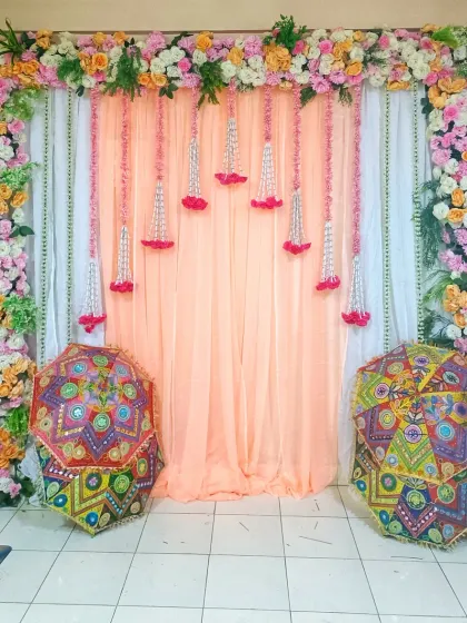 A beautiful backdrop for an at-home Haldi or Mehandi function. The setup includes a floral frame, hanging flower strings, and traditional Rajasthani umbrellas for a pop of color.