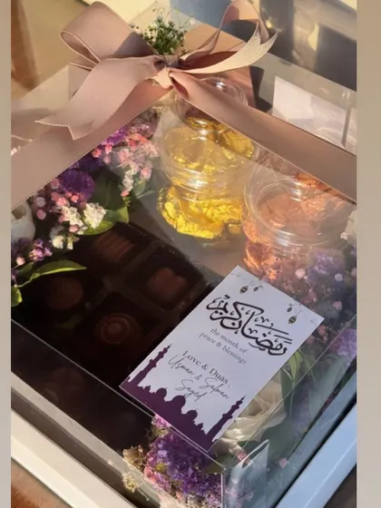 An elegant Eid hamper in a clear acrylic box. This gift includes jars of treats and a box of chocolates, surrounded by a beautiful arrangement of purple flowers and a custom "Ramadan Kareem" card.