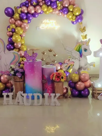 A unicorn party setup with a purple, pink, and gold color scheme, featuring a balloon arch, unicorn cutouts, and a light-up number "1".