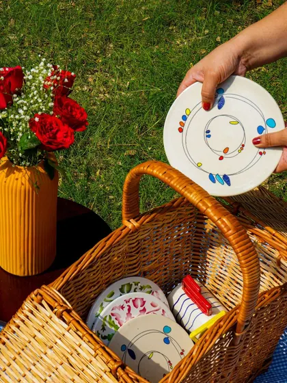 A lifestyle shot of a picnic scene, with a hand placing a plate into a wicker basket. This tells a story and shows the products in use.