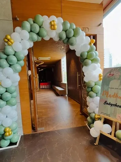 A simple and elegant balloon arch in sage green and white for a baby shower entrance, complemented by a personalized welcome board.
