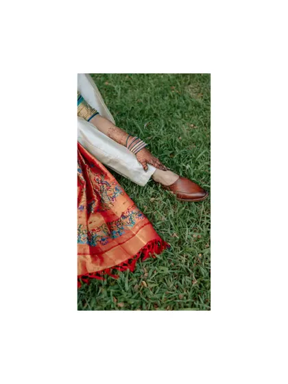 An artistic detail shot focusing on the bride's colorful saree and the groom's traditional jutti shoes, resting on the green grass.