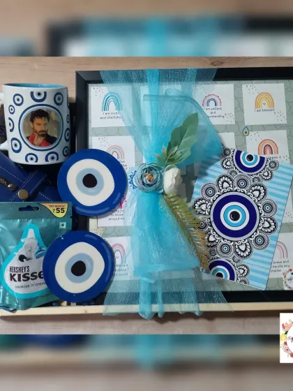 An evil eye themed hamper in a tray, containing a vision board, coasters, a personalized mug, a sunglasses case, and chocolates.
