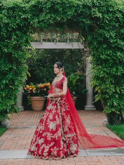 A poised and elegant portrait. The bride stands under a lush green archway, her red lehenga a beautiful contrast against the natural backdrop.