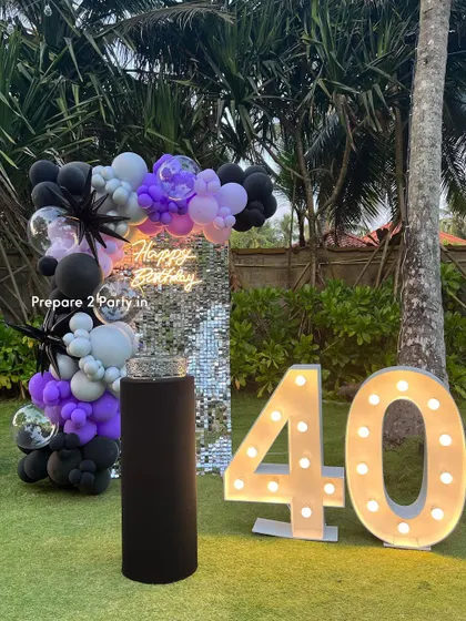 An outdoor 40th birthday celebration with a modern twist. This design includes a silver sequin wall, a balloon garland in black, purple, and silver, and large marquee numbers on the lawn.