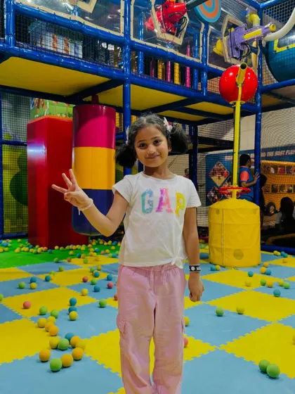 A girl flashes a peace sign in the middle of the ball pit area, surrounded by colorful balls and play structures.