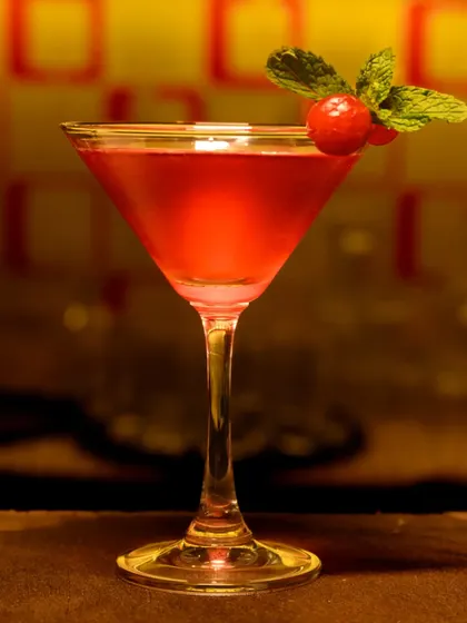 A vibrant red cocktail in a martini glass, garnished with a cherry and mint. The warm, ambient background of the bar adds to the sophisticated and inviting mood of the shot.