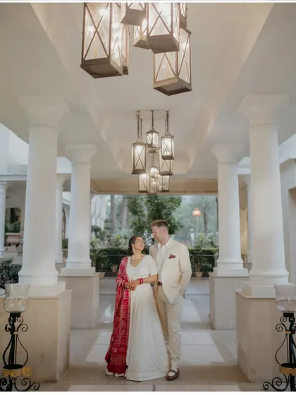 An elegant portrait of the couple in a beautifully lit corridor. The bride's red dupatta adds a stunning pop of color to their classic white reception outfits.