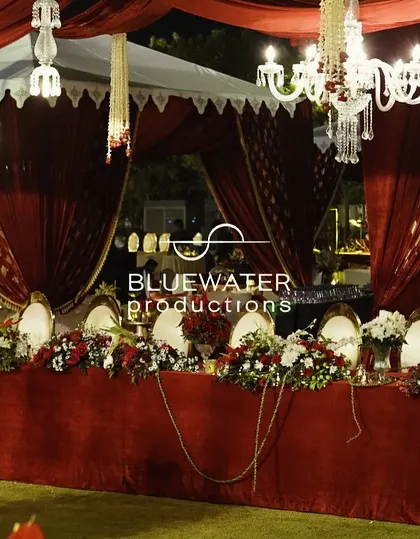 A long, communal dining table draped in red velvet and adorned with white and red floral arrangements, set under a canopy of chandeliers.