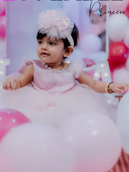 The little princess of the party. This photo captures the birthday girl sitting amidst a sea of pink and white balloons, looking adorable in her party dress and matching headband.