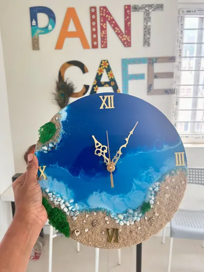 A classic beach resin clock with deep blue water, a sandy shore, and gold Roman numerals, held up against our studio backdrop.